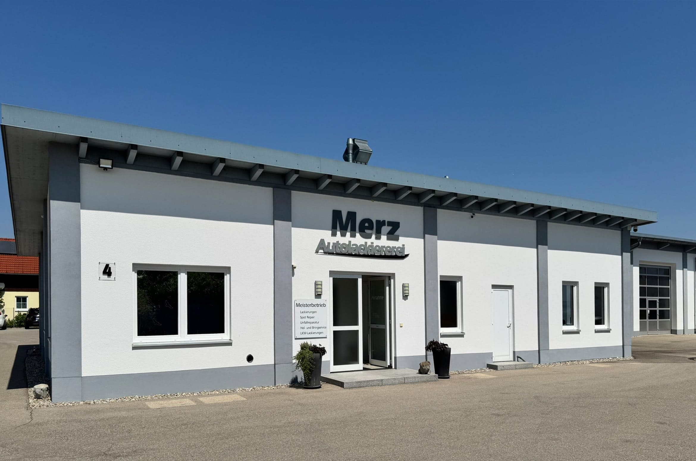 Leichte seitliche Ansicht von Firma Autolackiererei Merz mit Logo Merz Autolackiererei über dem Eingang - Gebäude weiß mit grauen Elementen